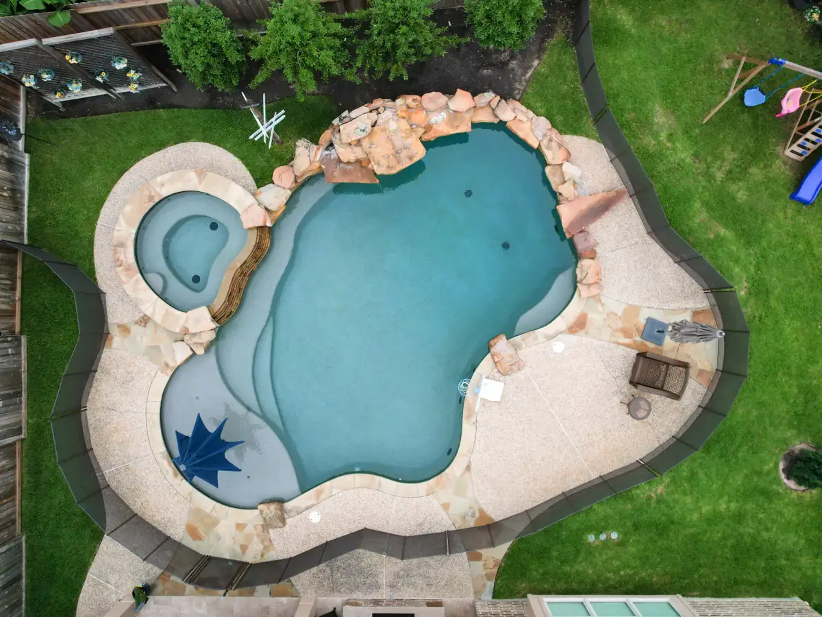 Drone view of a freeform pool with an attached round spa, star mosaic on the steps and a boulder waterfall
