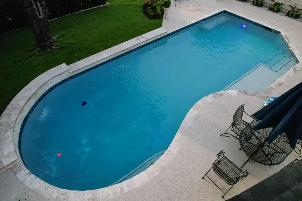 Drone view of a geometric pool with a travertine deck, sun shelf, lawn and patio chairs