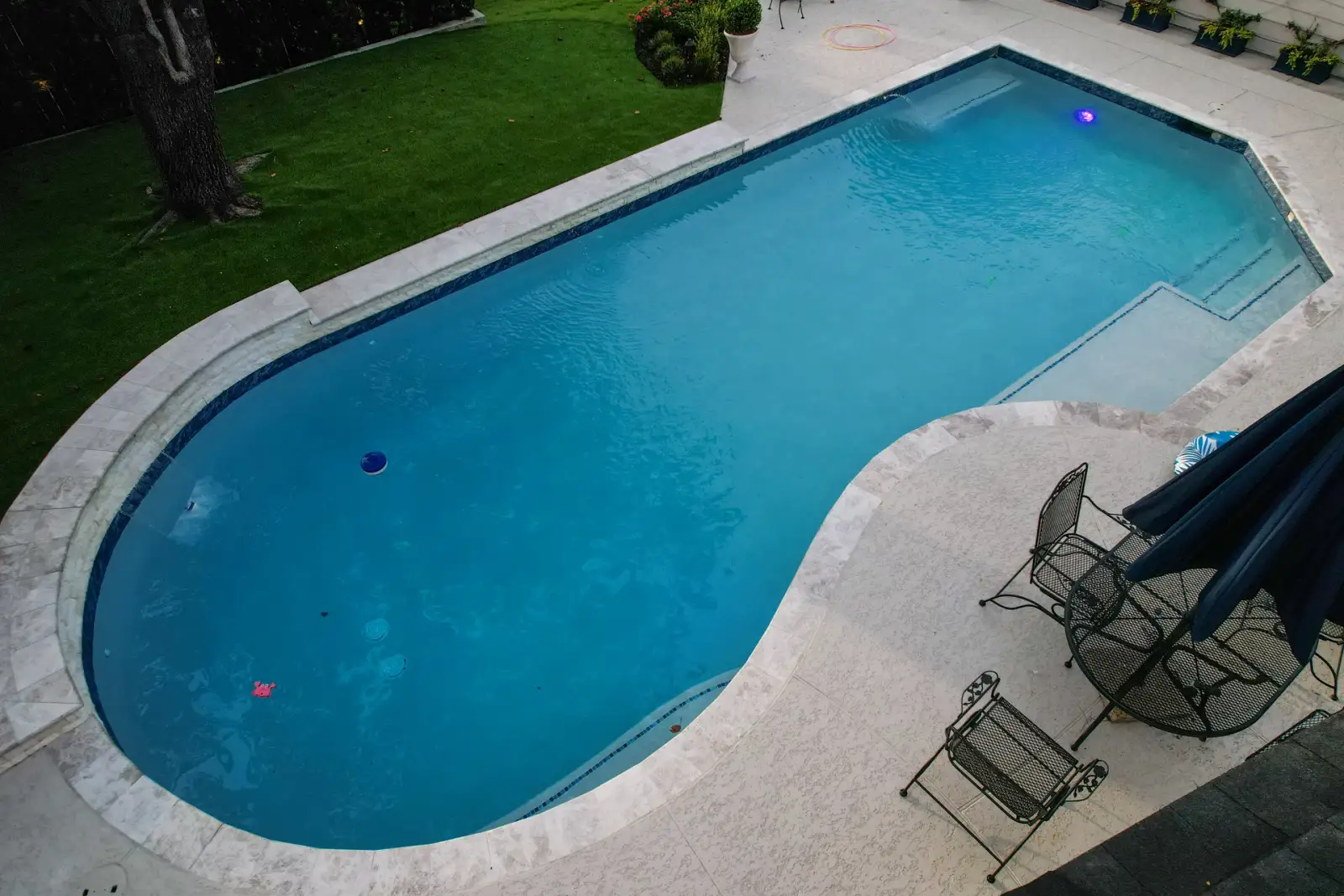 Drone view of a geometric pool with a travertine deck, lawn and patio seating