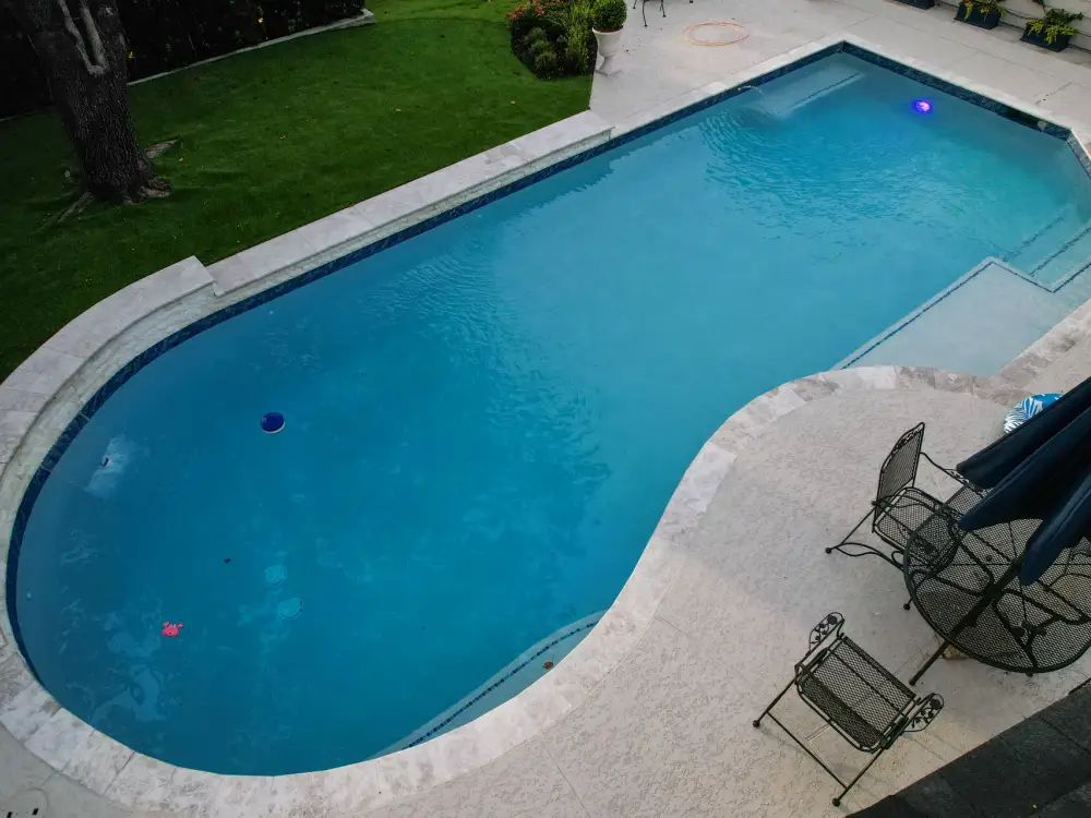 Drone view of a geometric pool with a travertine deck, sun shelf, lawn and patio chairs