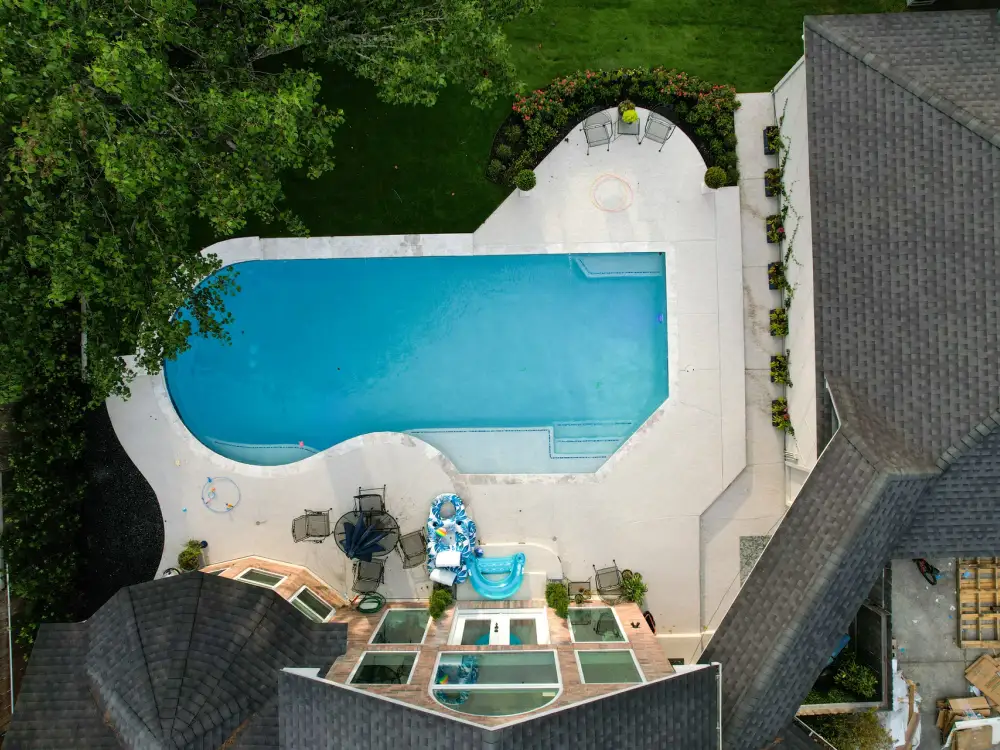 Overhead drone view of a rectangular pool with a curved end, white deck and patio seating beside the house
