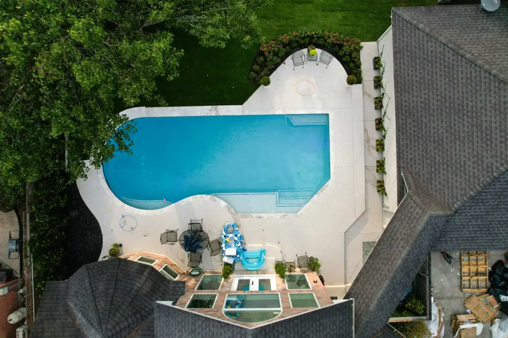Overhead drone view of a rectangular pool with a curved end, white deck and patio seating beside the house