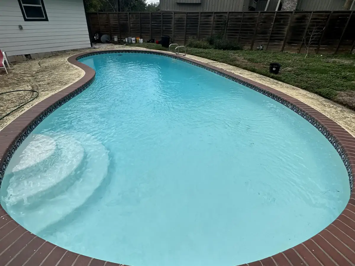 Kidney-shaped pool refilled with clear blue water after a white replaster, with red brick coping and a timber house behind