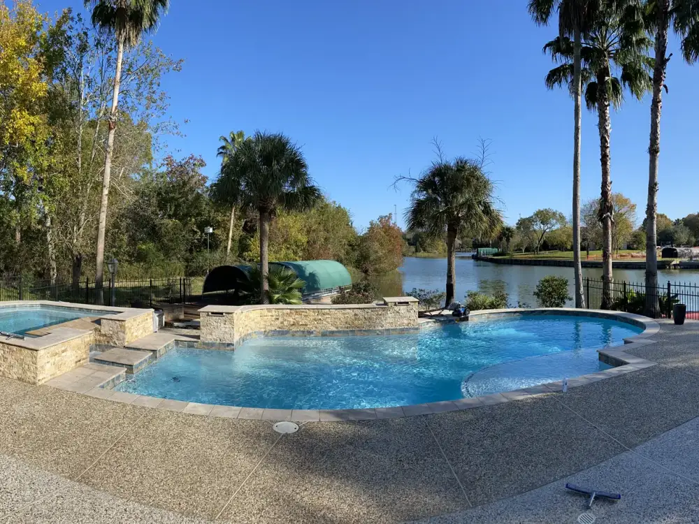 Wide panorama of a lakeside freeform pool with a raised spa, palm trees and an exposed-aggregate deck