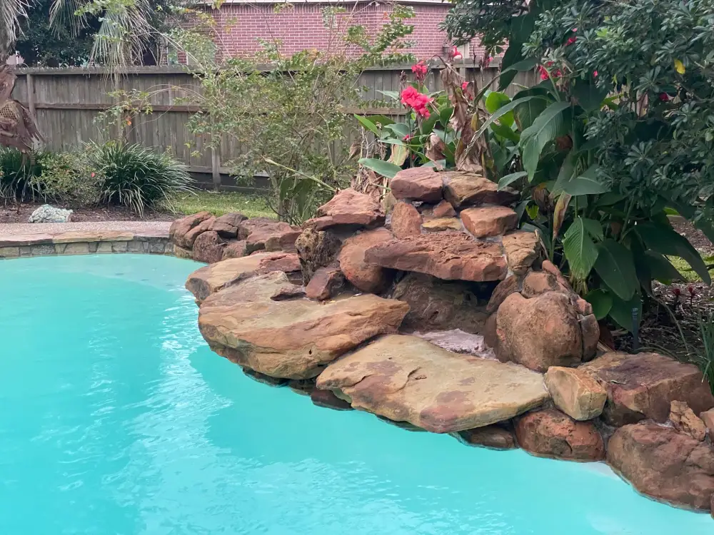 Stacked boulder waterfall at the edge of a pool with bright blue water and tropical planting