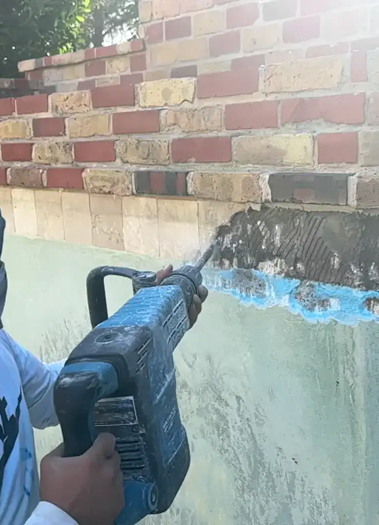 Aloha crew member chipping old blue waterline tile off a drained pool wall below brick coping