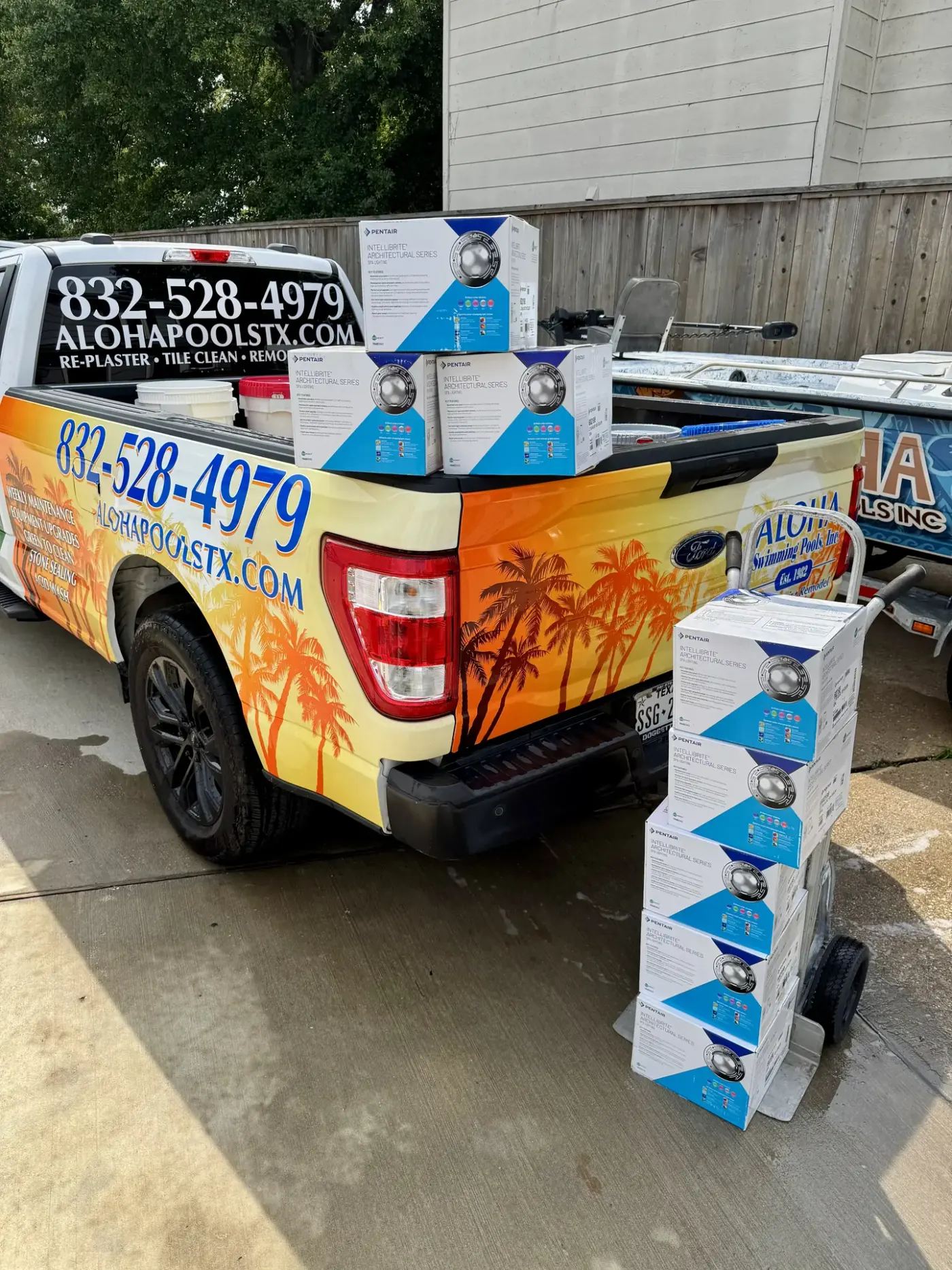 Aloha-branded pickup with LED pool light boxes stacked on the tailgate and a hand truck