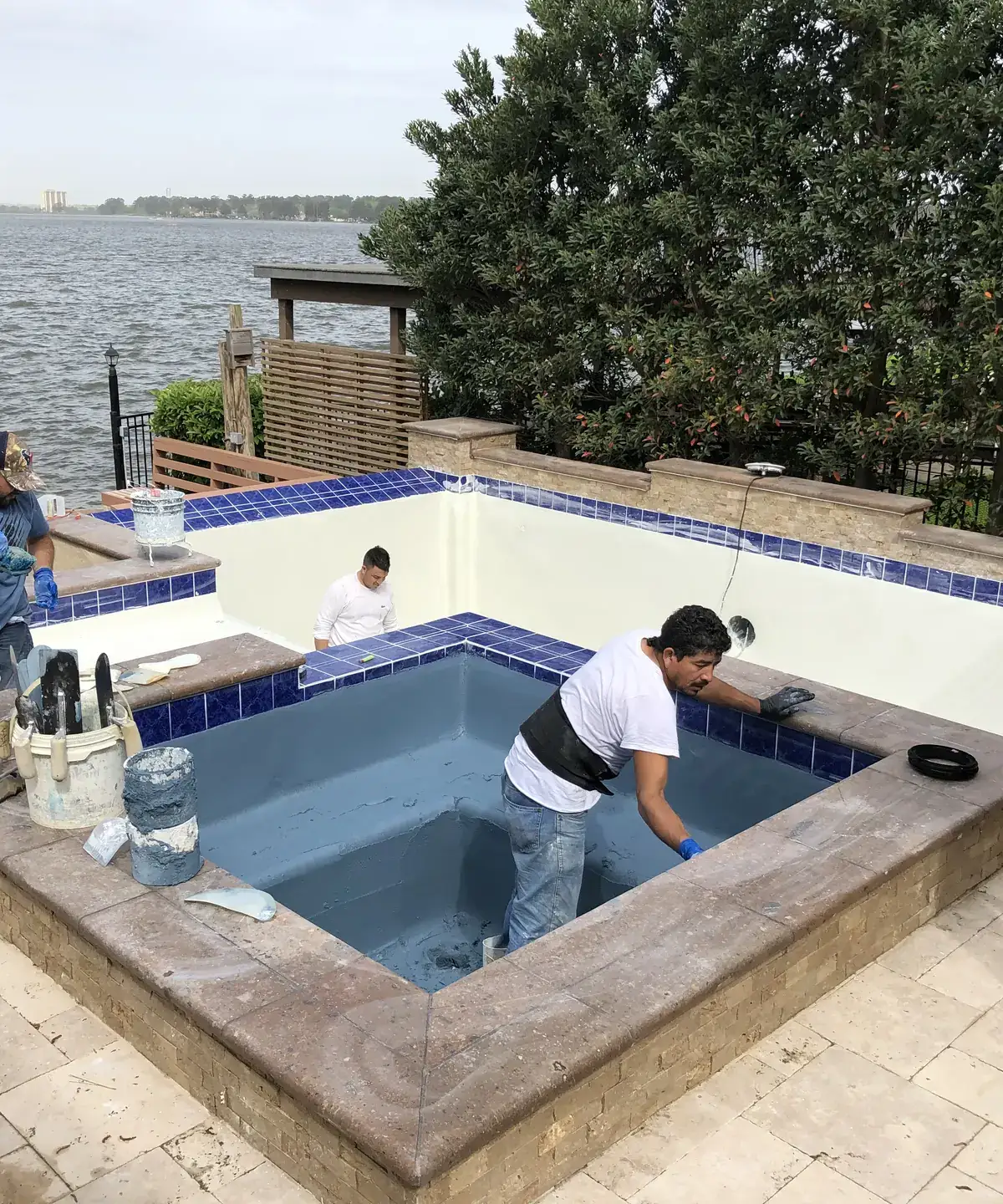 Aloha crew replastering a drained raised spa with blue waterline tile beside a lake