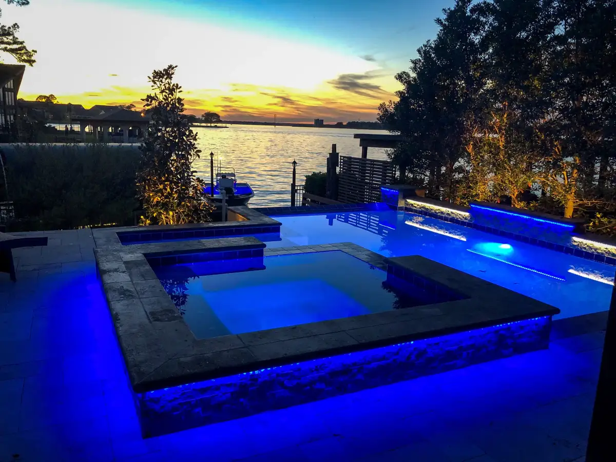 Waterfront pool and raised square spa glowing blue with LED lighting under a sunset sky